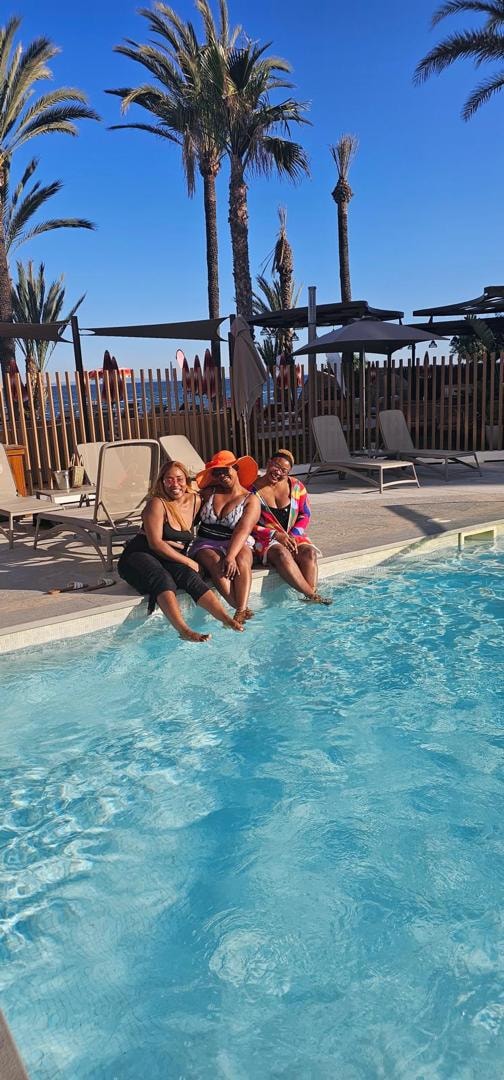 Three friends in sun hats sitting side by side at the edge of a turquoise resort pool lined with palm trees