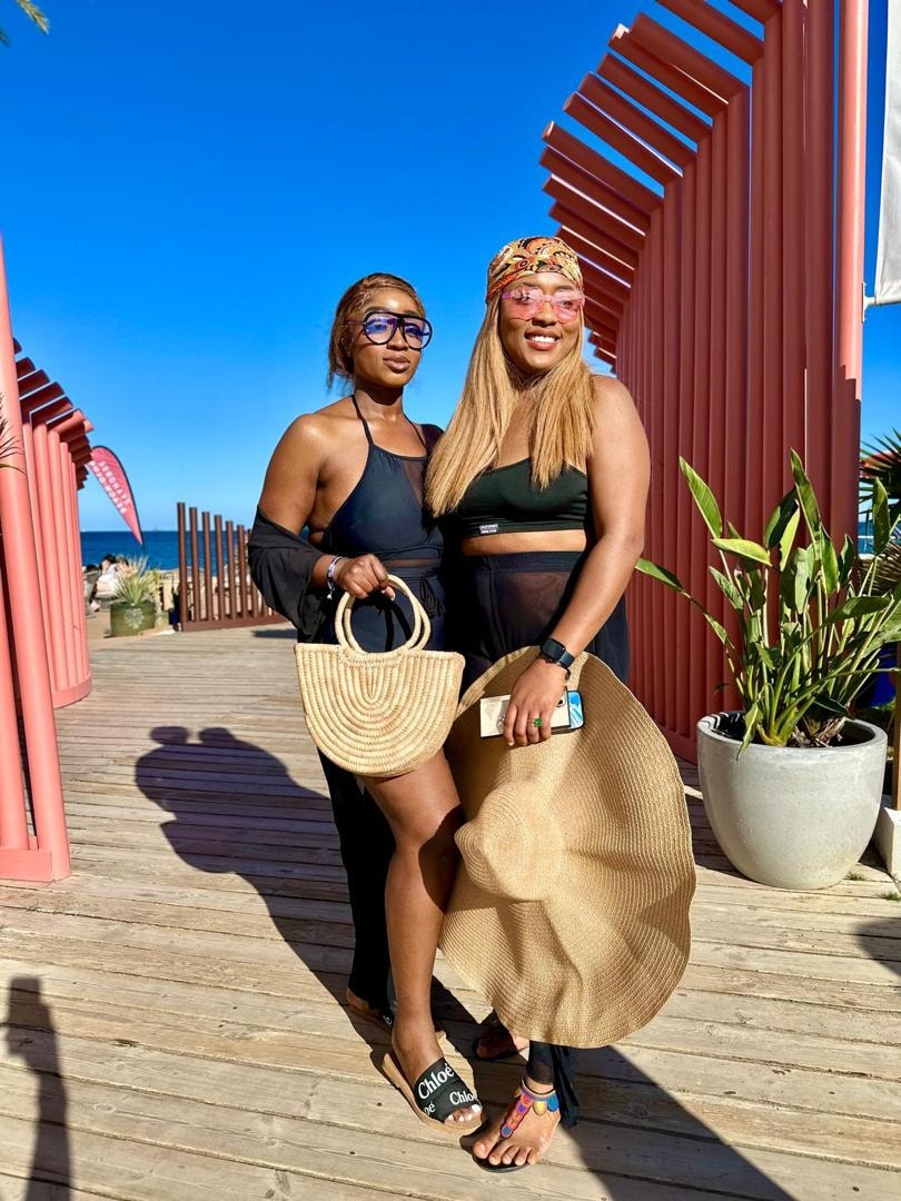 Two travellers in black beach outfits with woven bags posing on a boardwalk beneath a coral-red pergola by the sea